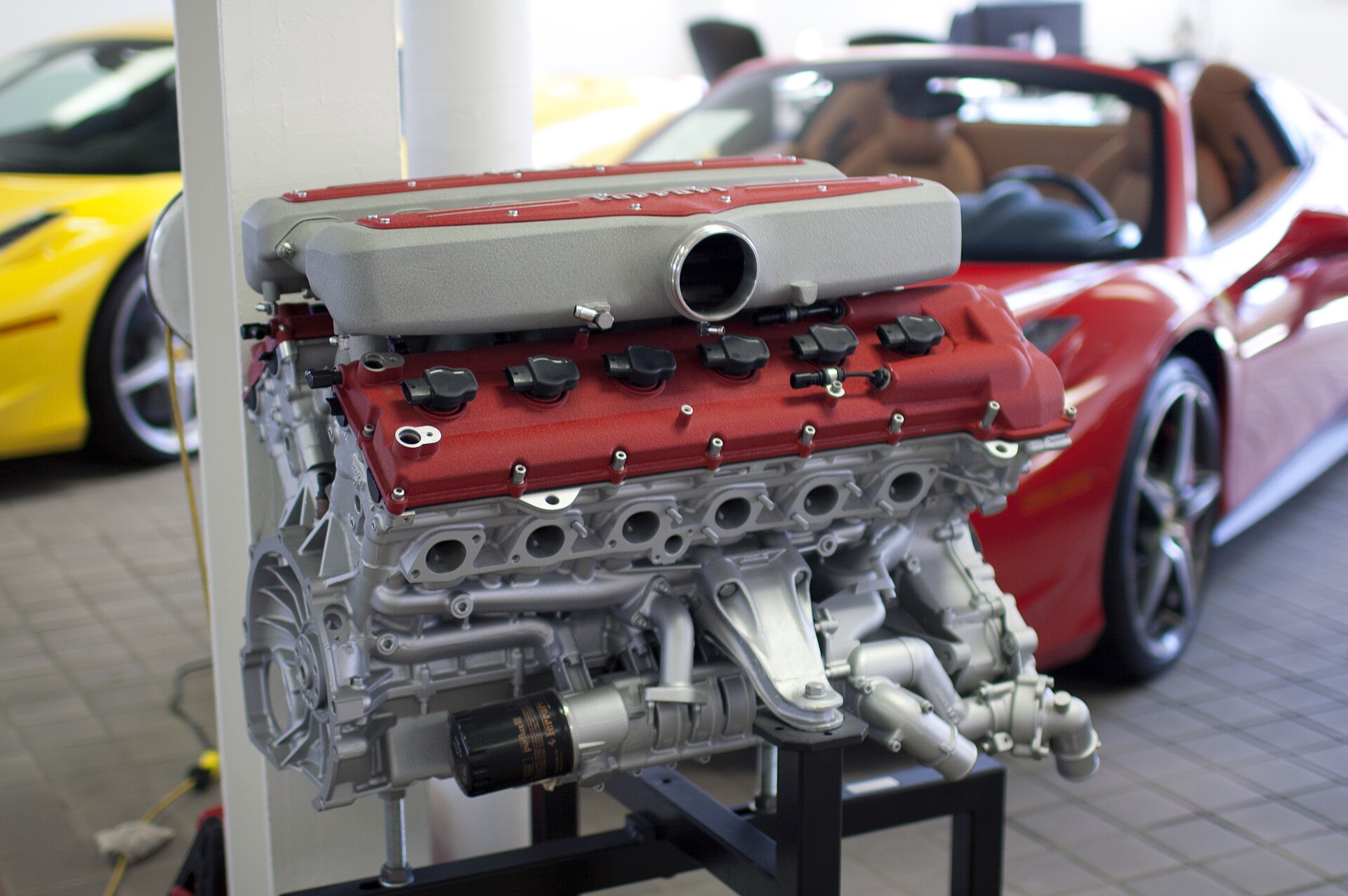 A Ferrari V12 engine on a stand in a showroom, cam covers in red crackle finish.